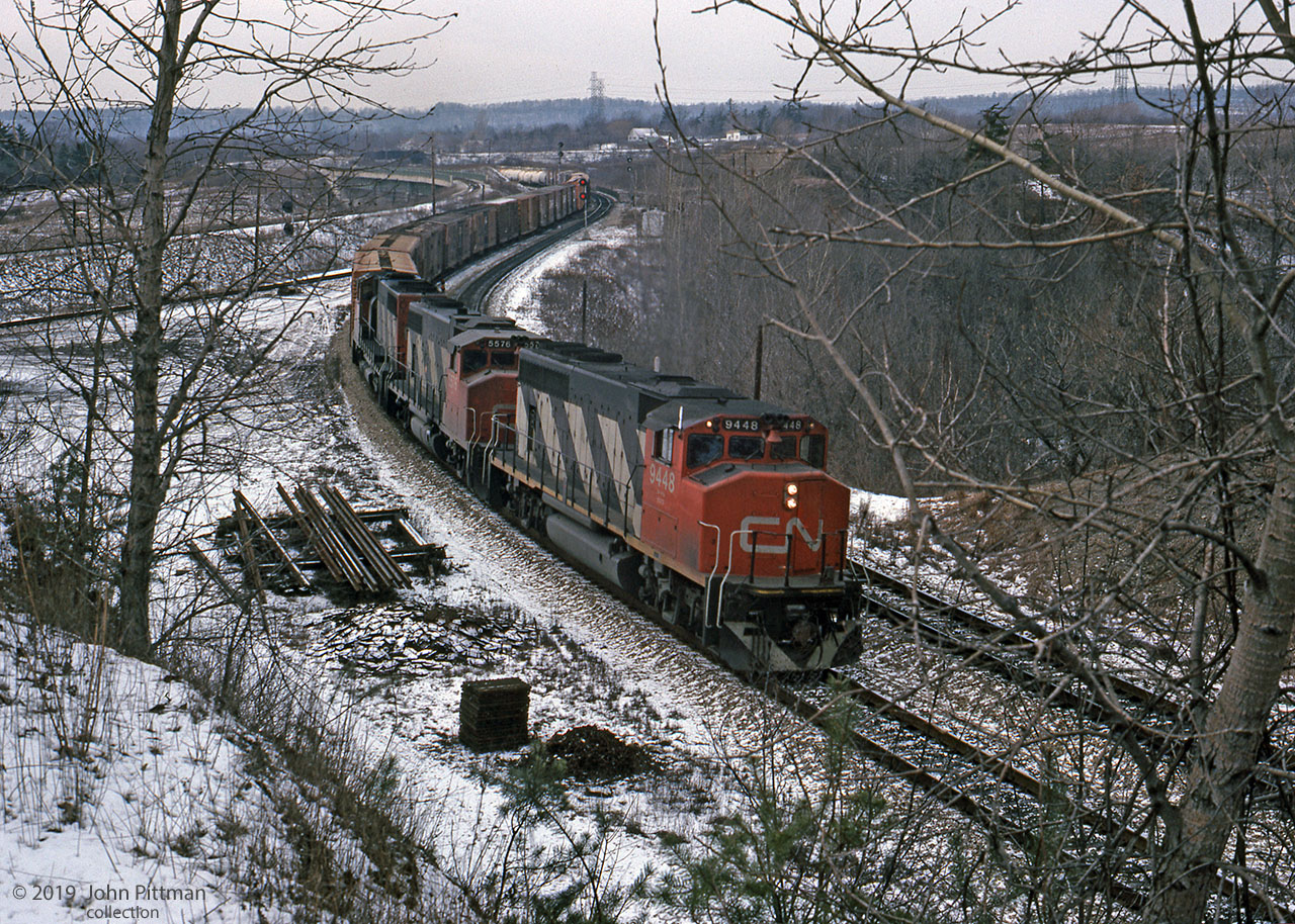 Four axle GMD locomotives CN 9448 (GP40-2LW), CN 5576 (GP38-2W), and CN 9540 lead an Extra east through CN Hamilton West at fractional mileage on the Dundas sub, near the bottom of the grade from Copetown. 
The gap in the trees where the photographer stood has grown in, but good views remain steps ahead. In 2019 the far part of the CP "S" curve and bridge over CN are obscured, jointed rails are not stockpiled for this main line, and westbound Dundas sub signals are down at Bayview.
CN 5576 was renumbered to CN 4776 in 1988; it was seen switching railcars in Aldershot Yard on several dates in 2017 in the current www.cn.ca paint scheme.